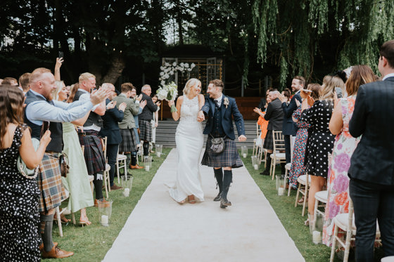Bride and groom smile together as they walk back down the aisle at The Venue, Eskmills while their guests clap